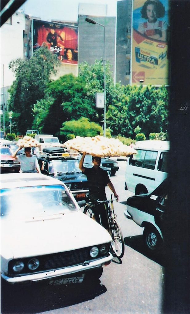 2 teenagers carrying loafs of bread on top of a wooden shelf while riding a bicycle in traffic